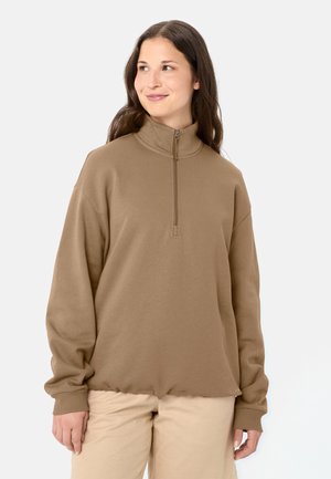 Frau mit langen braunen Haaren, die einen braunen Pullover mit Viertelreißverschluss und beiger Hose trägt, schaut leicht lächelnd nach links.