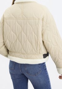 Beżowa pikowana kurtka z kołnierzem i mankietami z kożucha, z dopasowaną talią i detalem wentylacyjnym z tyłu, zestawiona z dżinsami.