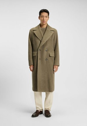 Olive green double-breasted coat with herringbone texture, wide lapels, and two front pockets, paired with cream trousers and dark loafers.