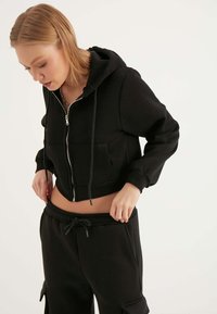 Felpa nera corta con zip, cappuccio regolabile con lacci, tasca a marsupio e orlo a costine, abbinata a pantaloni della tuta neri coordinati.