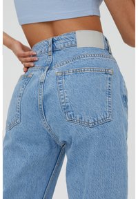 Jeans denim azzurri chiari con vita alta, caratterizzati da una superficie testurizzata, cuciture delle tasche laterali e un'etichetta sul retro di colore grigio liscio.