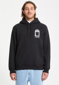 Haut de hoodie noir en tissu doux avec une poche avant, présentant un design graphique blanc sur la poitrine et des cordons de serrage sur la capuche.