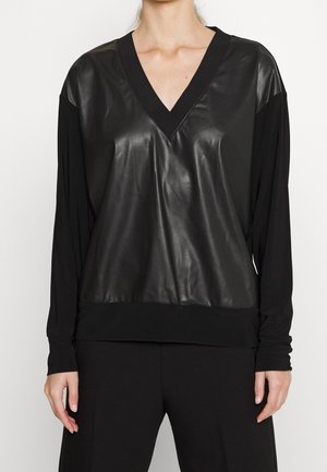 Sort langærmet V-hals top med skinnende læderlignende frontpanel og matte ærmer, båret af en person mod en ensfarvet baggrund.