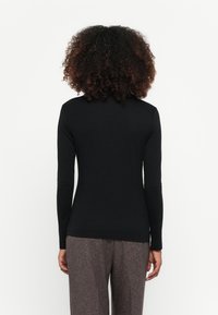 Frau mit lockigem Haar, die ein enganliegendes schwarzes Langarmoberteil und graue, strukturierte Hosen trägt, von hinten vor einem weißen Hintergrund gezeigt.