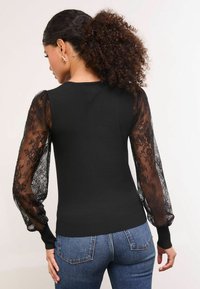 Fekete felső, áttetsző csipkés puffujjakkal, egyszínű testtel és bordázott mandzsettákkal. Kék farmernadrággal párosítva. A ujjakon részletes virágmintás csipketervezés található.