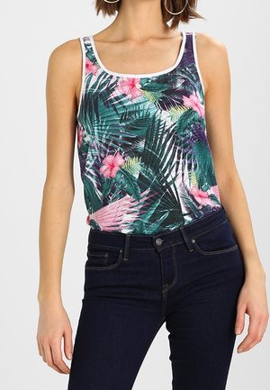 Nainen, jolla on yllään hihaton tropiikin kukkakuvioinen toppi ja tummansiniset farkut, seisoo käsivarsien ollessa rentona sivuilla.