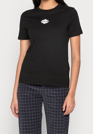 Kobieta w czarnej koszulce Diesel z białym logo na piersi i ciemnoszarych spodniach w kratę, stojąca na jasnym tle.