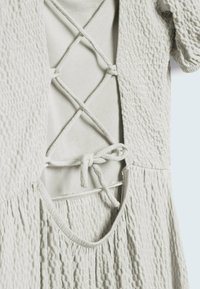 Gros plan sur une robe texturée beige clair avec des liens croisés lacés dans le dos et des manches courtes bouffantes.