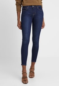 Mujer vestida con jeans ajustados azul oscuro y sandalias de tacón de estampado animal marrón, de pie contra un fondo claro y liso.