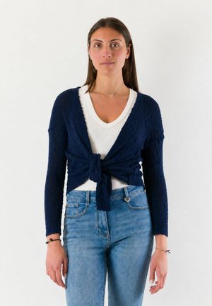 Giovane donna in piedi, indossa un cardigan blu navy a manica lunga annodato sul davanti sopra una maglietta bianca a scollo a V e jeans blu.