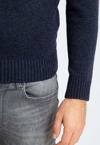 Oberkörper eines Mannes, der einen dunkelblauen gestrickten Pullover und graue Jeans trägt, wobei eine sichtbare Hand neben der Tasche ruht.