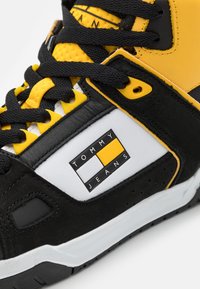 Tenisi negri și galbeni, de tip high-top, cu un finisaj din piele texturată și textil, având un patch alb cu logo și accente galbene.