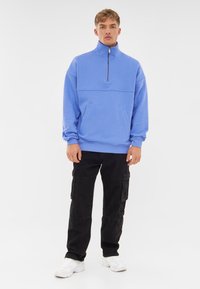 Lichtblauwe half-zip sweatshirt met twee voorzakken, geribde manchetten en zoom, gecombineerd met zwarte cargobroeken en witte sneakers.