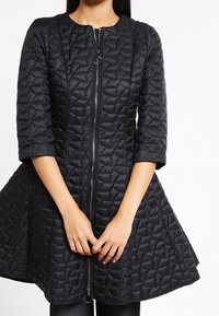 Chaqueta acolchada negra con mangas tres cuartos y un bajo acampanado, con cierre frontal con detalle de anilla.