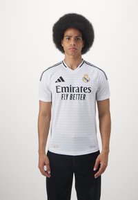 Maglia da calcio bianca con accenti neri, caratterizzata dal logo "Emirates Fly Better" e dallo stemma del Real Madrid. Texture liscia, maniche corte, design a V.