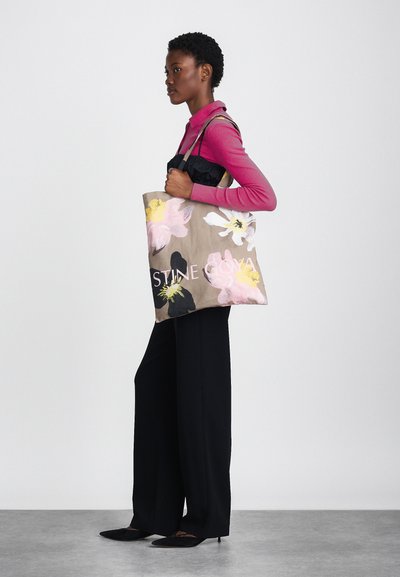 Canvas tote bag med blommönster i rosa, vitt och gult på en beige bakgrund; har en logotyp och kraftiga handtag.