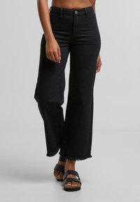 Kvinde iført sort højtaljede jeans med vide ben og frynser ved kanten samt sorte platformsandaler, stående på en grå overflade.