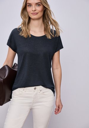 Kobieta o blond włosach do ramion, ubrana w ciemnoszarą koszulkę i białe spodnie, trzymająca dużą brązową skórzaną torbę.