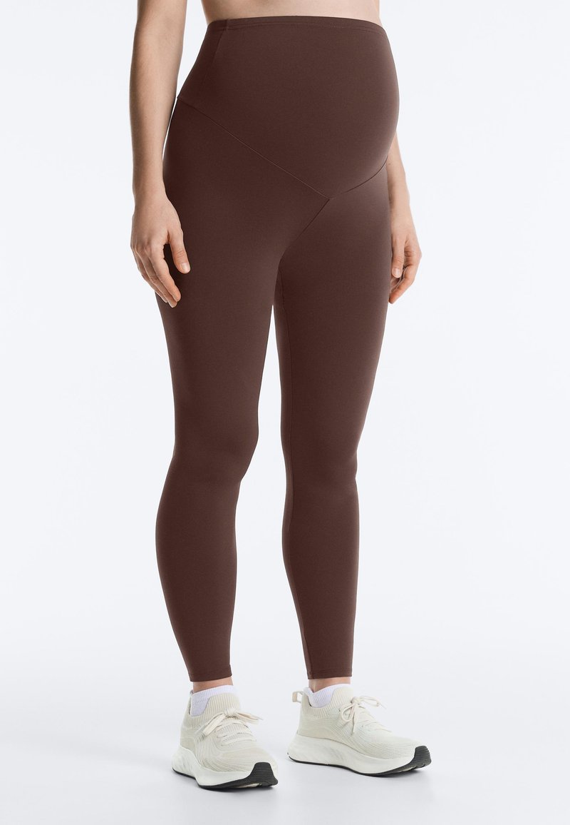 Personne enceinte portant un legging de maternité marron et des baskets blanches, debout devant un fond uni.