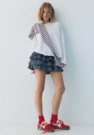 Ung kvinde iført hvid langærmet top med en stribet skjorte hængende over skulderen, lagdelt ternet mininederdel, røde sokker og røde sneakers.