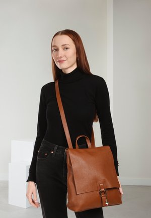 Brown leather backpack with a smooth texture, a flap closure, and adjustable shoulder strap. Model wearing black turtleneck and jeans.