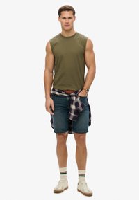 Canotta verde oliva, pantaloni shorts di jeans, camicia a quadri annodata in vita, sneakers bianche e marroni e calze a righe.