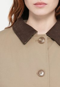 Casaco bege com um colarinho de veludo cotelê castanho-escuro e fecho de botão. O tecido parece suave, com uma textura sutil e um design ajustado.