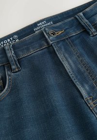 Sötétkék farmernadrág, a derékrész közeli nézete. Jellemzője a látható varrás, egy gomb és egy címke, amelyen a "Comfort Stretch Next Performance" felirat olvasható.