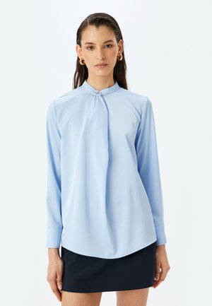Camicia a maniche lunghe di colore azzurro chiaro con colletto alto e dettaglio a bottoni sul collo. Tessuto liscio con orlo curvo, abbinata a una gonna scura.