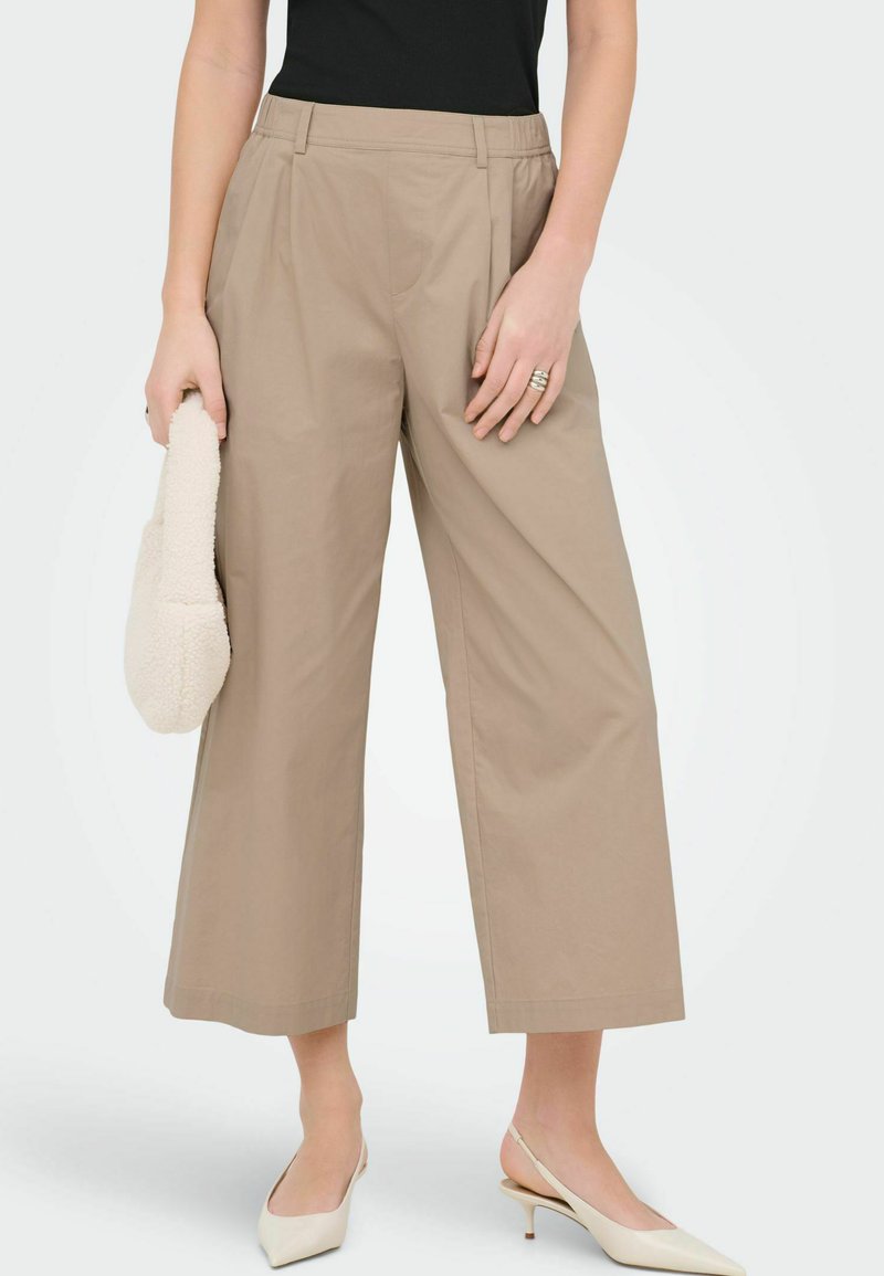 Kvinde iført beige bukser med vide ben og korte ben, sort top, hvide slingback-hæle, holder en hvid tekstureret håndtaske og har ringe på fingrene.