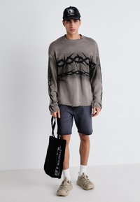 Pull en tricot gris surdimensionné avec un motif abstrait noir, shorts gris, baskets beiges et sac fourre-tout noir. Le modèle se tient devant un fond uni.