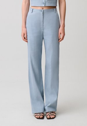 Donna che indossa pantaloni a vita alta e gamba ampia color azzurro chiaro e sandali neri aperti con fiocchi, in piedi davanti a uno sfondo semplice.