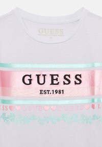 Bela organska bombažna majica s središnjim logotipom "GUESS" v črni barvi, okrašena s pastelno roza in turkiznimi črtami ter cvetličnimi vzorci.