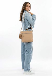 Beige leather crossbody bag with a zip closure and tassel detail. Soft texture and simple design, worn over a light denim jacket.