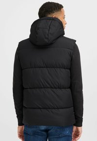 Schwarze Pufferweste mit Kapuze, ausgestattet mit einem gesteppten Design, elastischen Armausschnitten und einer glatten Textur, getragen über einem schwarzen Langarmoberteil.