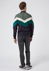 Sudadera con patrones de chevron en verde, crema y azul marino; tejido de mezcla de algodón. Combinada con pantalones gris oscuro y zapatillas casuales.