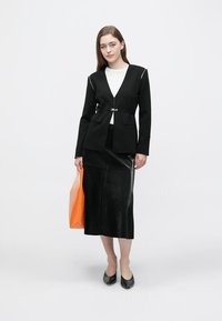 Svart skräddarsydd kavaj med vita kantdetaljer, kombinerad med en svart läder-midikjol och en orange handväska. Modellen bär svarta ballerinaskor.