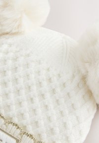 Fehér kötött sapka mintás textúrával, tetején két bolyhos pomponnal és az alján fémes díszítéssel.