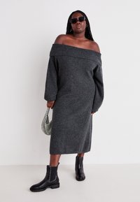 Robe tricotée grise, à manches longues et décolletée sur les épaules, mi-mollet. Associée à des bottines noires à clous et à un petit sac à perles.
