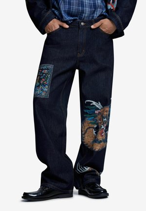 Jean en denim foncé à jambes larges, arborant des patchs brodés colorés de dragons et de fleurs, porté avec des chaussures noires et les mains dans les poches.