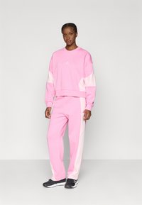 Rozā sporta tērps, kas izgatavots no mīkstas auduma, ar baltām akcents uz piedurknēm un sāniem. Ietver mazu logotipu uz krūtīm un brīvu piegriezumu.