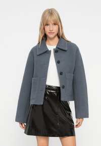 Donna bionda che indossa una giacca grigia a bottoni con texture sopra una maglietta bianca e una gonna nera lucida, in piedi davanti a uno sfondo uniforme.