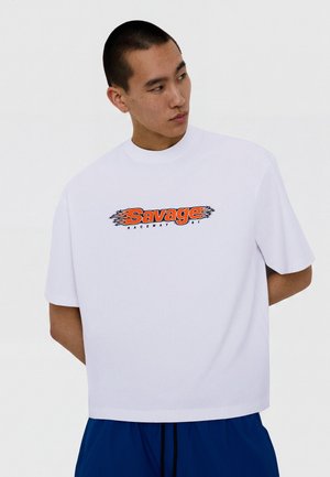 Jeune homme aux cheveux courts portant un t-shirt blanc oversize avec le logo flamme "Savage Raceway 9.1" en orange et noir, et un pantalon bleu.