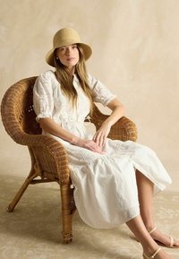 Weißes Augelet-Kleid mit Puffärmeln und Kordelzug in der Taille, kombiniert mit einem Strohhut, sitzend in einem geflochtenen Korbstuhl. Neutraler Hintergrund.