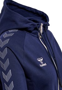 Marineblauwe hoodie met een rits aan de voorkant, een capuchon met trekkoord en een wit chevronpatroon langs de mouw. Geborduurd logo op de borst.
