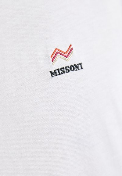 Valge T-särk Missoni logoga; omaduseks punased, roosad ja oranžid sakilised tikandid musta teksti kohal. Pehme kangas ja sile tekstuur.