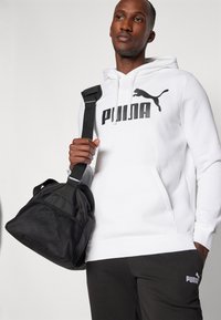 Hvid hættetrøje med sort logo, parret med en sort mesh- og stofduffelbag. Modellen holder tasken med en sort justerbar rem.