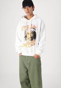 Sweatshirt branco com gráficos "Thug Life" e "California Love". Tecido suave, cordão, punhos canelados. Usado com calças largas de algodão verdes.