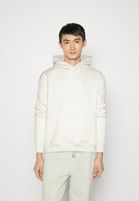Sudadera blanca con bolsillo delantero y capucha con cordón, confeccionada en una tela suave y con un logo sutil grabado en la parte delantera. Combinada con pantalones claros.