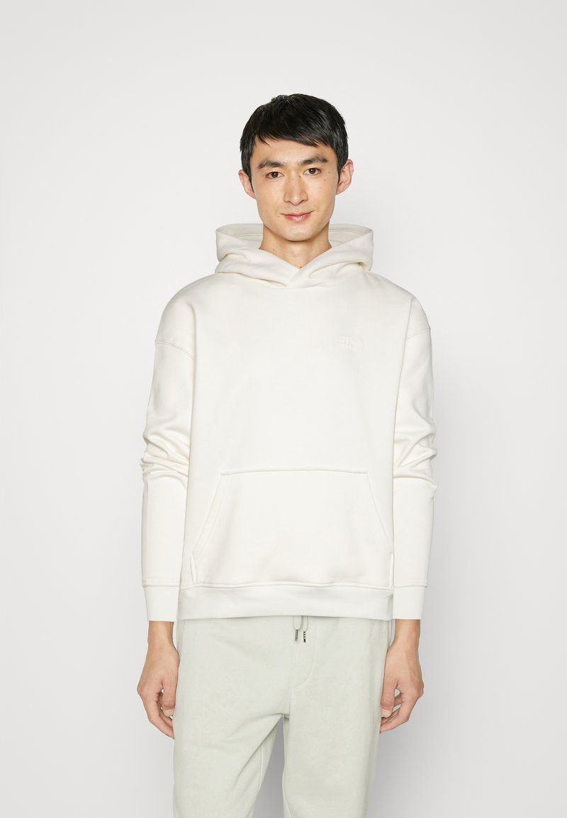 Sudadera blanca con bolsillo delantero y capucha con cordón, confeccionada en una tela suave y con un logo sutil grabado en la parte delantera. Combinada con pantalones claros.